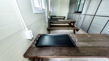A row of wooden tables with a black tray on one, a white electrical outlet on the wall, and a window with blinds.