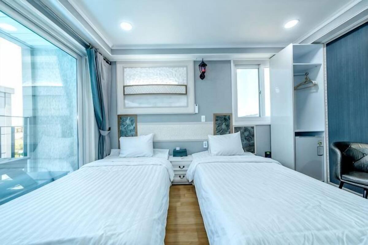 A hotel room with two beds, a large window, a wardrobe, and a small desk.