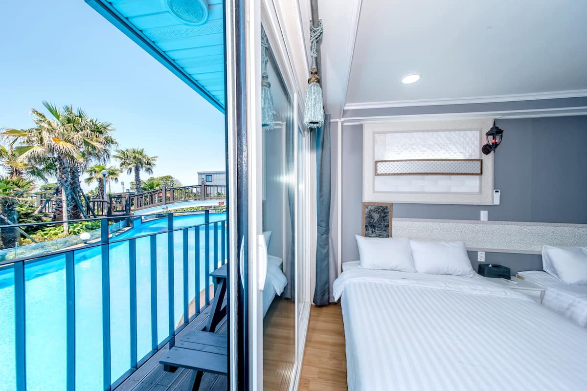 A balcony with a pool, a bed with white bedding, and a view of palm trees.