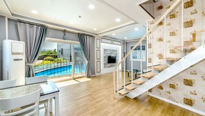 Ett modernt hotellrum med trägolv, en matplats med bord och stolar, en trappa med en vägg som har ett mönster och utsikt över en swimmingpool genom stora fönster.