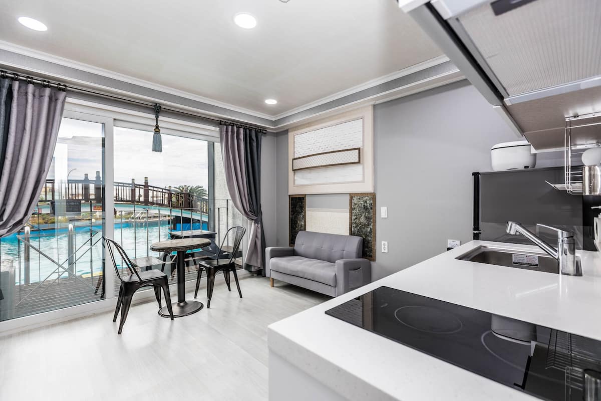 A modern hotel room with a dining area, a gray sofa, a kitchen with a built-in stove, and a view of an indoor pool.