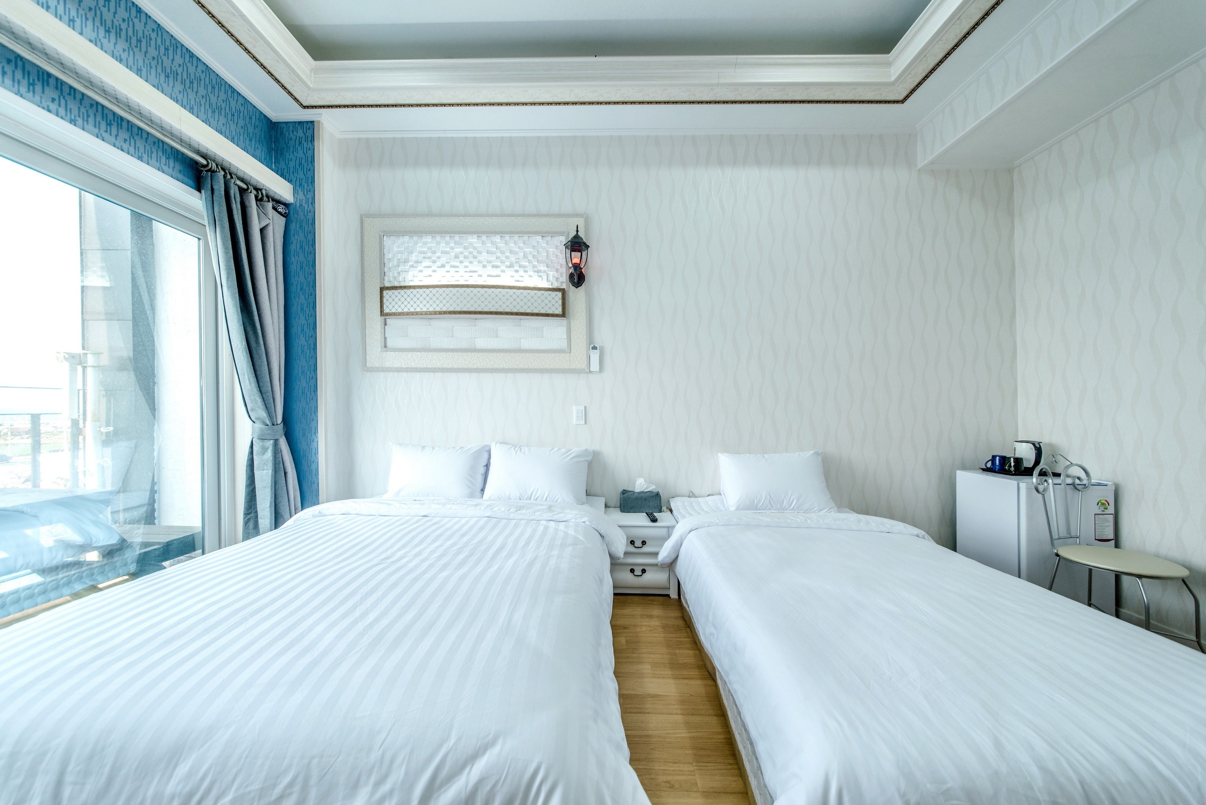A hotel room with two beds, a small desk, a chair, a window with curtains, and a wall-mounted lamp.