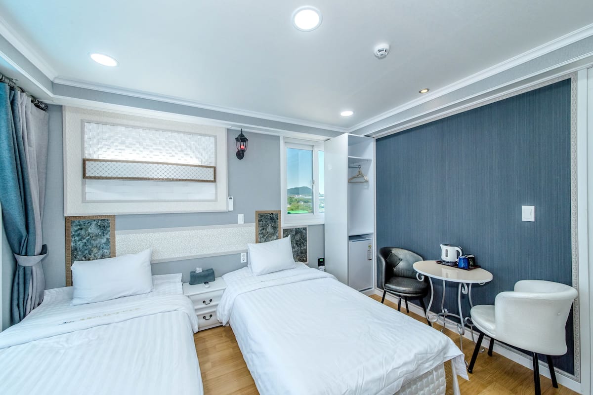A hotel room with two beds, a small table, a chair, and a view of the outdoors.