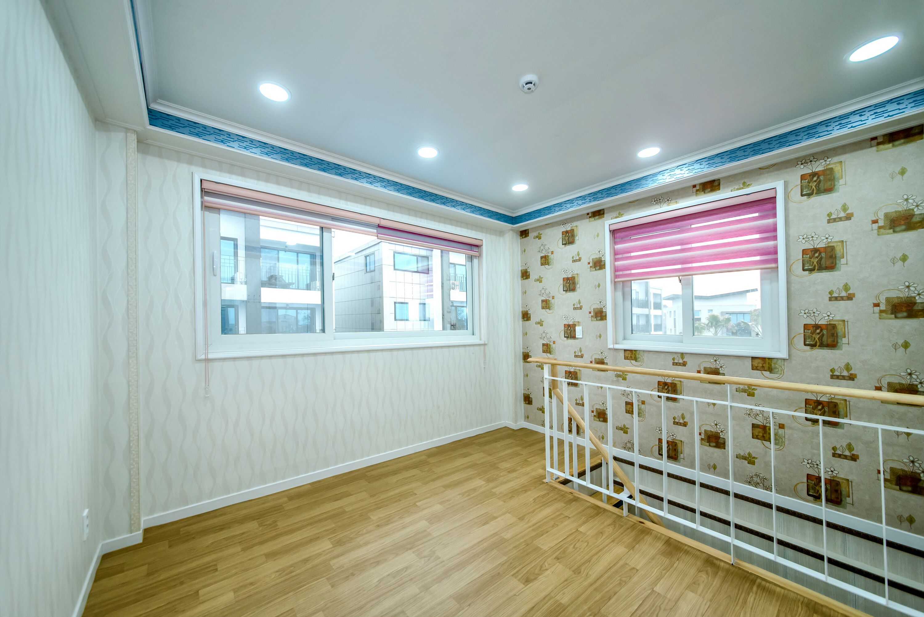 Una habitación con suelo de madera, paredes blancas y papel tapiz estampado. Hay dos ventanas con persianas, una barandilla blanca y un techo con iluminación empotrada.