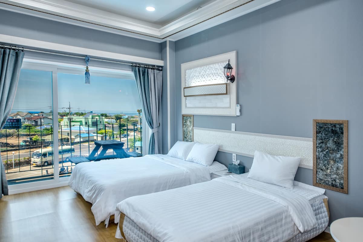 A hotel room with two beds, a large window with a view of the city, a small table, and a wall-mounted lamp.