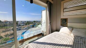 Un dormitorio con un ventanal grande, una cama con ropa de cama blanca y vistas a un paisaje urbano y una masa de agua.