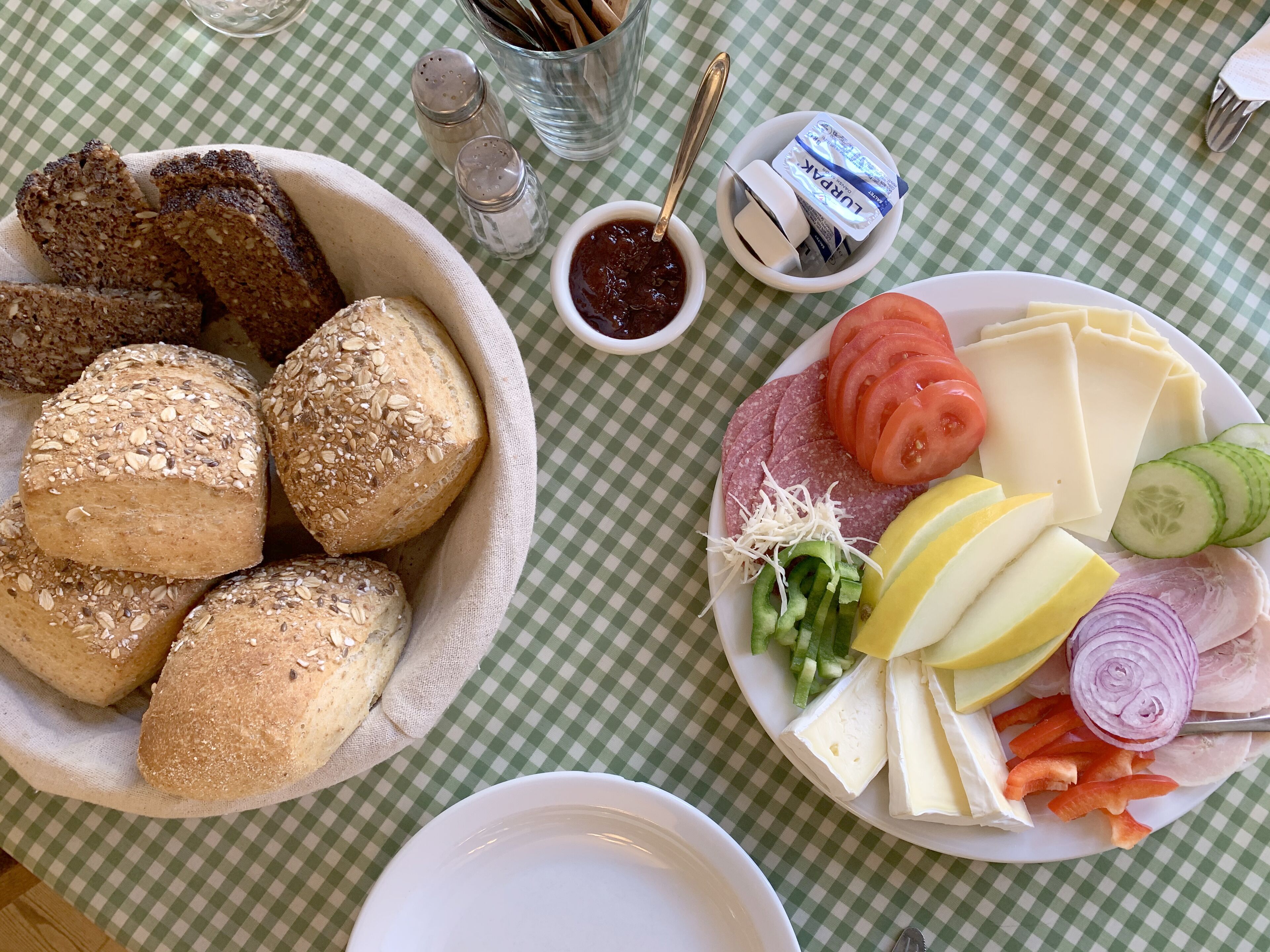 A table with a checkered tablecloth, a bowl of seeded bread, a plate with cheese, vegetables, and sliced meat.