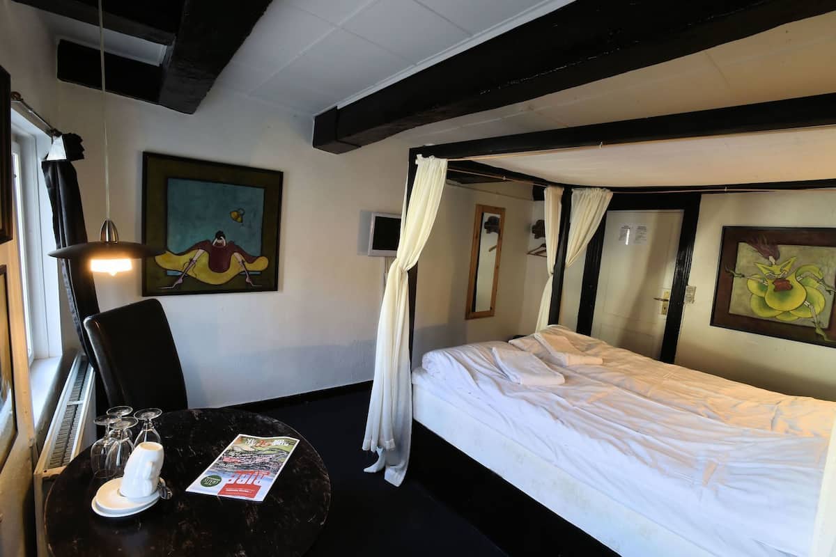 A hotel room with a bed, a chair, a table with a magazine, a painting, and a piano.