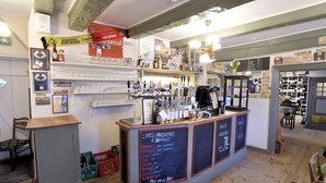 Un espace bar avec un tableau noir affichant le menu des boissons et leurs prix. On y trouve des étagères remplies de verres et de bouteilles, ainsi qu’un panneau indiquant la diffusion d’un événement de la Ligue des champions.