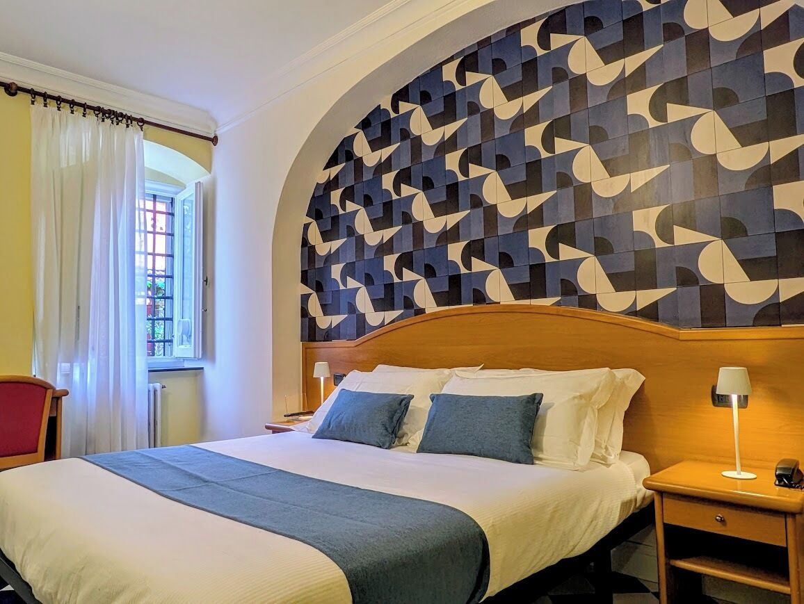 A hotel room with a bed, a nightstand, a lamp, a chair, and a window with curtains.
