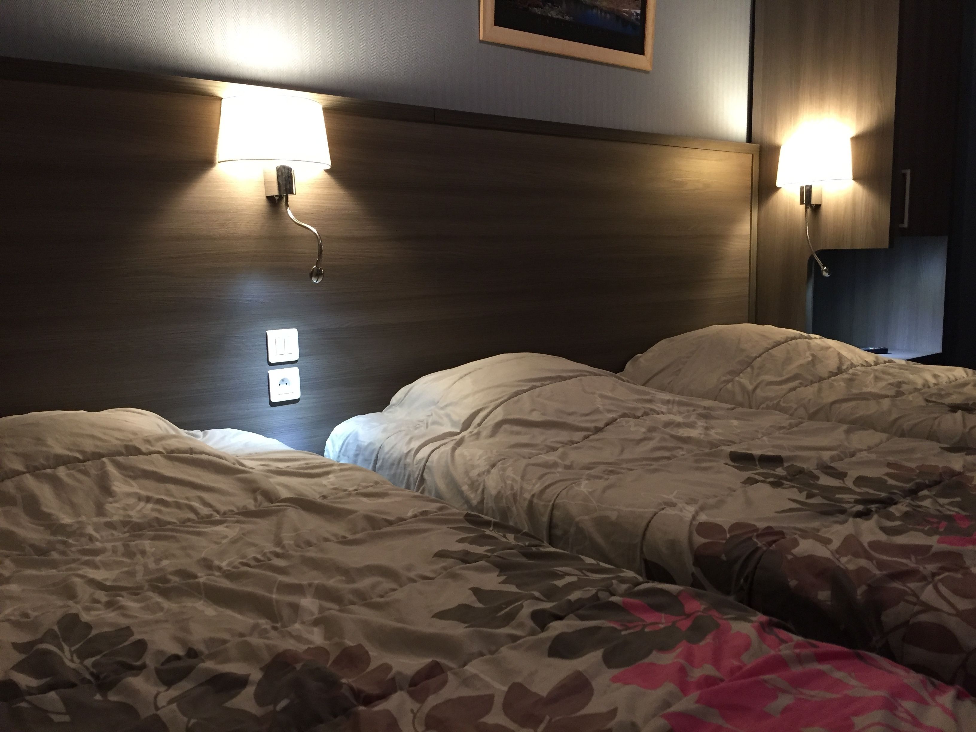 A hotel room with two beds, each with a headboard and bedside lamps. There are two wall-mounted light switches and a framed picture above the headboard.