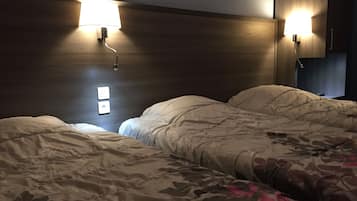 A hotel room with two beds, each with a headboard and bedside lamps. There are two wall-mounted light switches and a framed picture above the headboard.