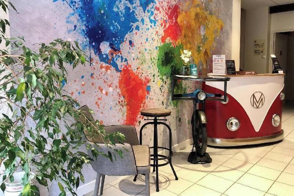 Un espace d’accueil agrémenté d’un mur décoré d’une œuvre abstraite aux couleurs vives, d’un comptoir de style vintage, d’un tabouret de bar et d’une plante en pot.