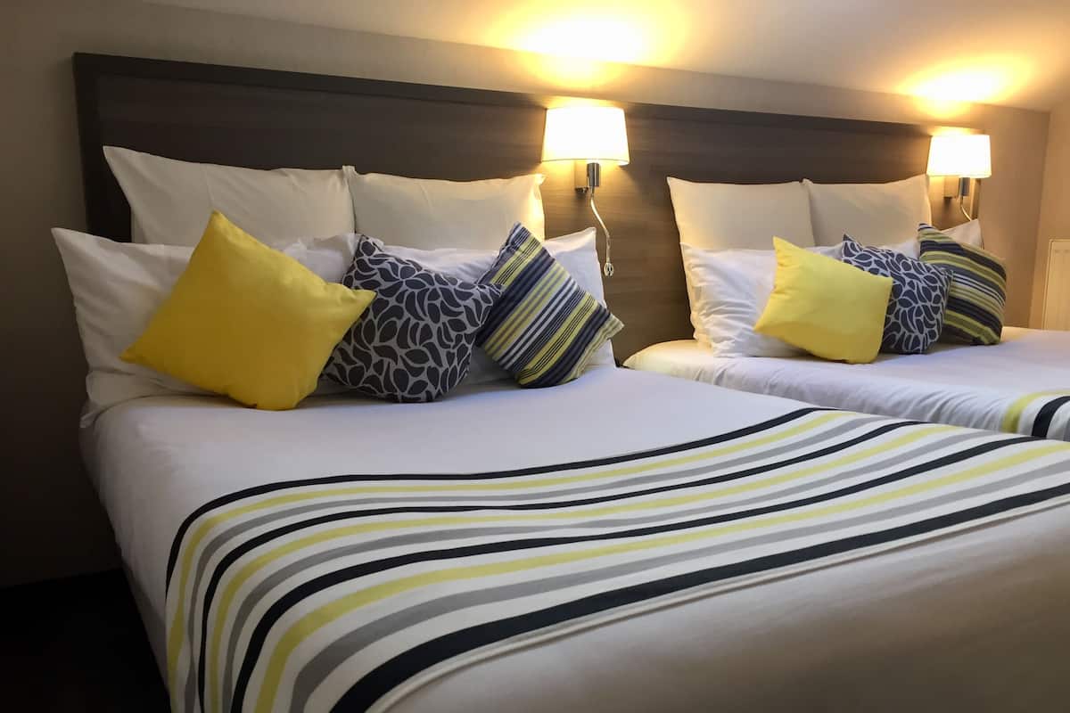 Une chambre d’hôtel avec deux lits, chacun doté de draps blancs et de couvertures à rayures. On y trouve plusieurs oreillers aux motifs et couleurs variés, notamment jaune et gris. La chambre est équipée d’une tête de lit en bois et de lampes fixées au mur pour l’éclairage.
