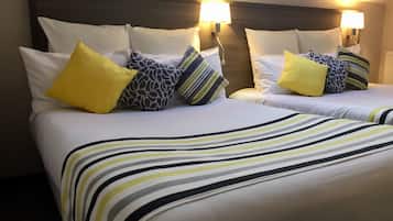 Ein Hotelzimmer mit zwei Betten, jeweils mit weißer Bettwäsche und gestreiften Decken. Mehrere Kissen in verschiedenen Mustern und Farben, darunter gelb und grau, sorgen für Komfort. Das Zimmer verfügt über ein Holz-Kopfteil und wandmontierte Lampen, die für Licht sorgen.