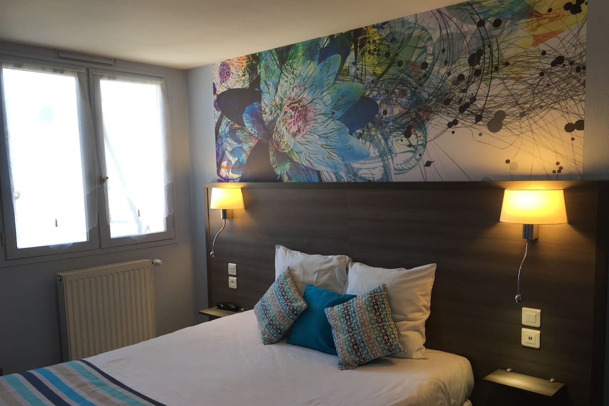 Une chambre d’hôtel avec un grand lit, deux lampes de chevet, un radiateur et un tableau abstrait aux couleurs vives accroché au mur.