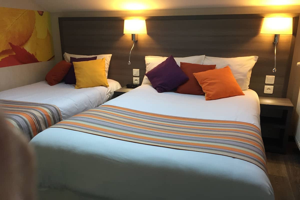 Une chambre d’hôtel avec deux lits, chacun doté de draps à rayures et de plusieurs oreillers de différentes couleurs. Des lampes murales éclairent la pièce, et un grand tableau abstrait orne le mur.