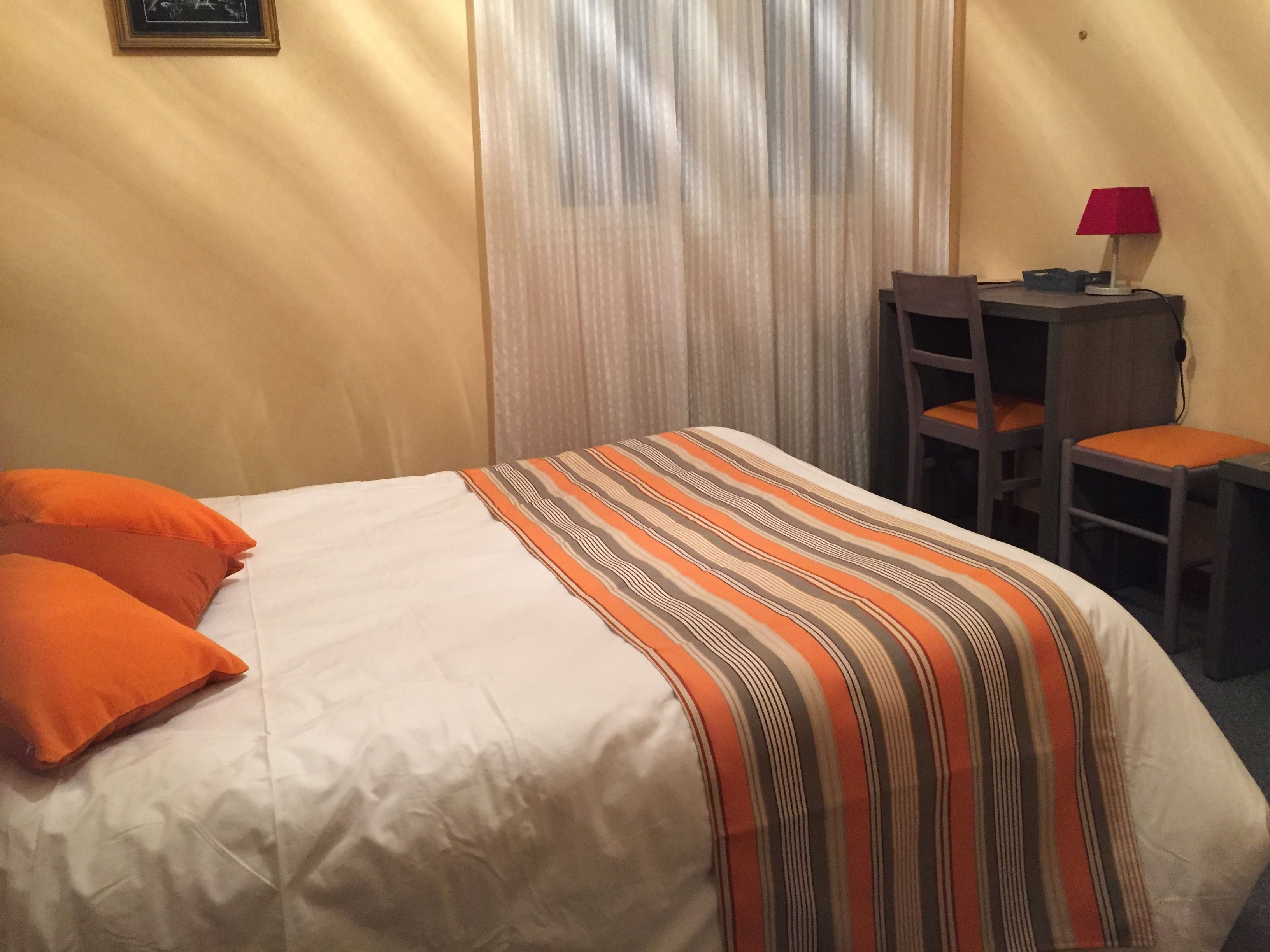 Une chambre d’hôtel avec un lit, deux oreillers orange, une couverture à rayures, un bureau avec une chaise et une lampe rouge.