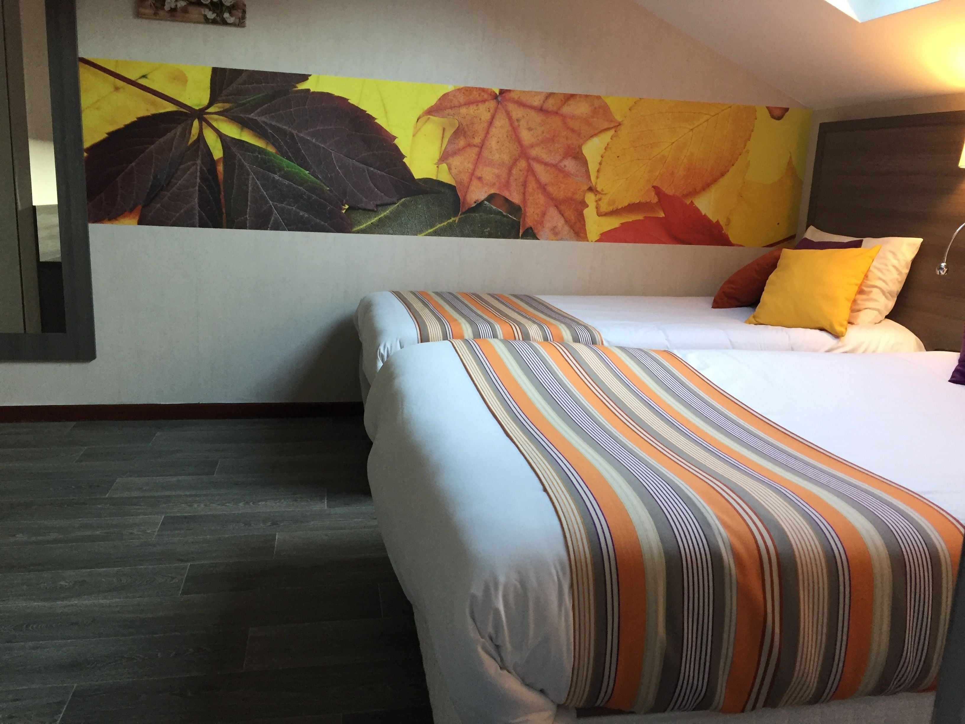 A hotel room with a bed, a striped pillow, a wall with autumn leaves, and a dark wooden floor.