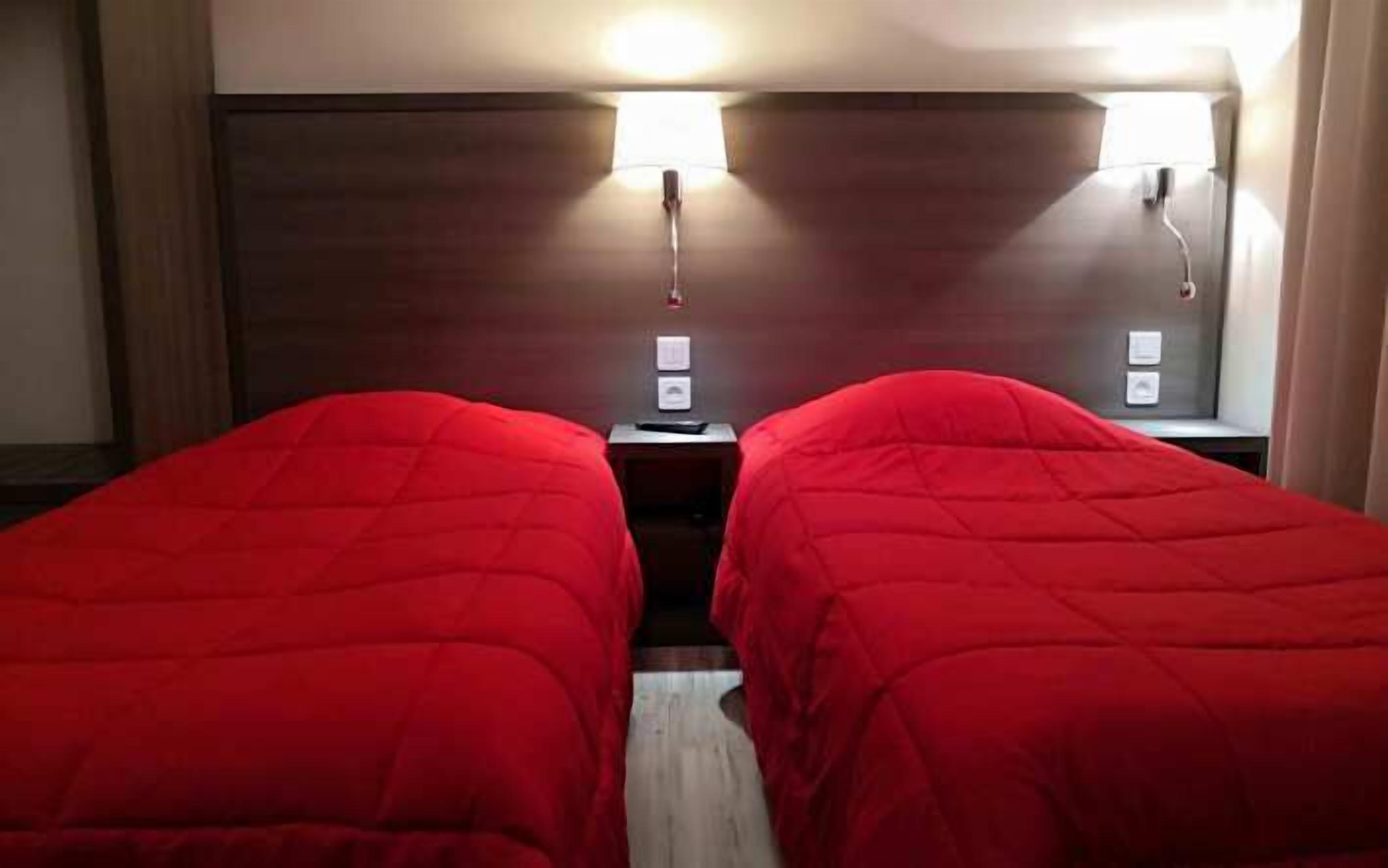 Une chambre d’hôtel avec deux lits simples, chacun recouvert d’une courtepointe rouge, dotée d’une tête de lit en bois et de deux lampes de chevet.