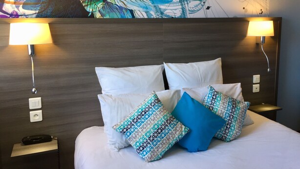 Une chambre d’hôtel avec un grand lit, du linge de lit blanc et des coussins à motifs. Deux lampes murales sont fixées de chaque côté du lit, et une prise de courant se trouve sur le mur.