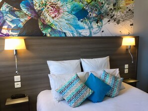 Habitación de hotel con una cama grande, ropa de cama blanca y almohadas estampadas. Hay dos lámparas de pared a cada lado de la cama, y un enchufe en la pared.