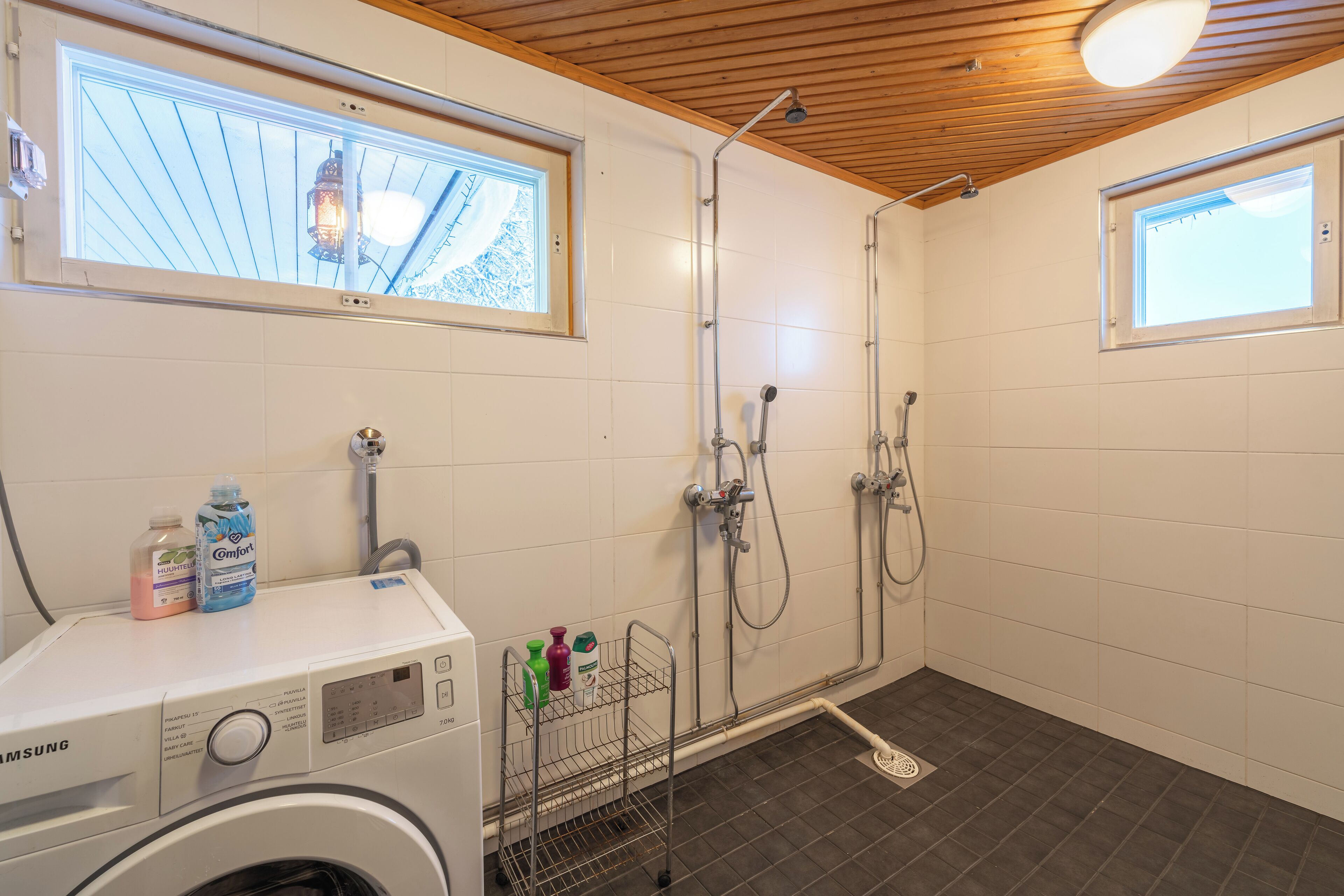 Une salle de bain compacte équipée d’un lave-linge, d’une douche avec plusieurs robinets et d’une fenêtre laissant entrer la lumière naturelle.