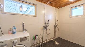 Une salle de bain compacte équipée d’un lave-linge, d’une douche avec plusieurs robinets et d’une fenêtre laissant entrer la lumière naturelle.
