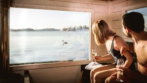 A cabin with a large window overlooking a lake, a swan, and a person holding a glass. - Almåsa Havshotell (Vasterhaninge)