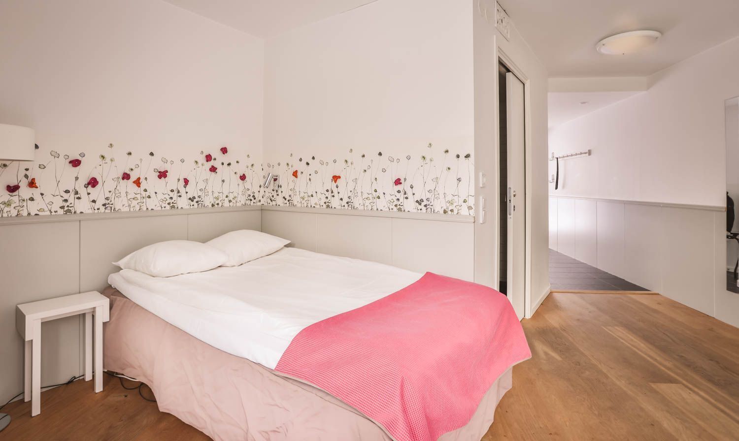 A bedroom with a bed, a nightstand, a pink blanket, and a wall with a floral design.