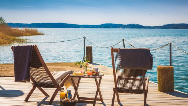 Two wooden lounge chairs with towels, a table with a drink and snacks, and a view of a lake with a distant shoreline. - Almåsa Havshotell (Vasterhaninge)