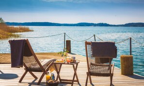Two wooden lounge chairs with towels, a table with a drink and snacks, and a view of a lake with a distant shoreline. - Almåsa Havshotell (Vasterhaninge)