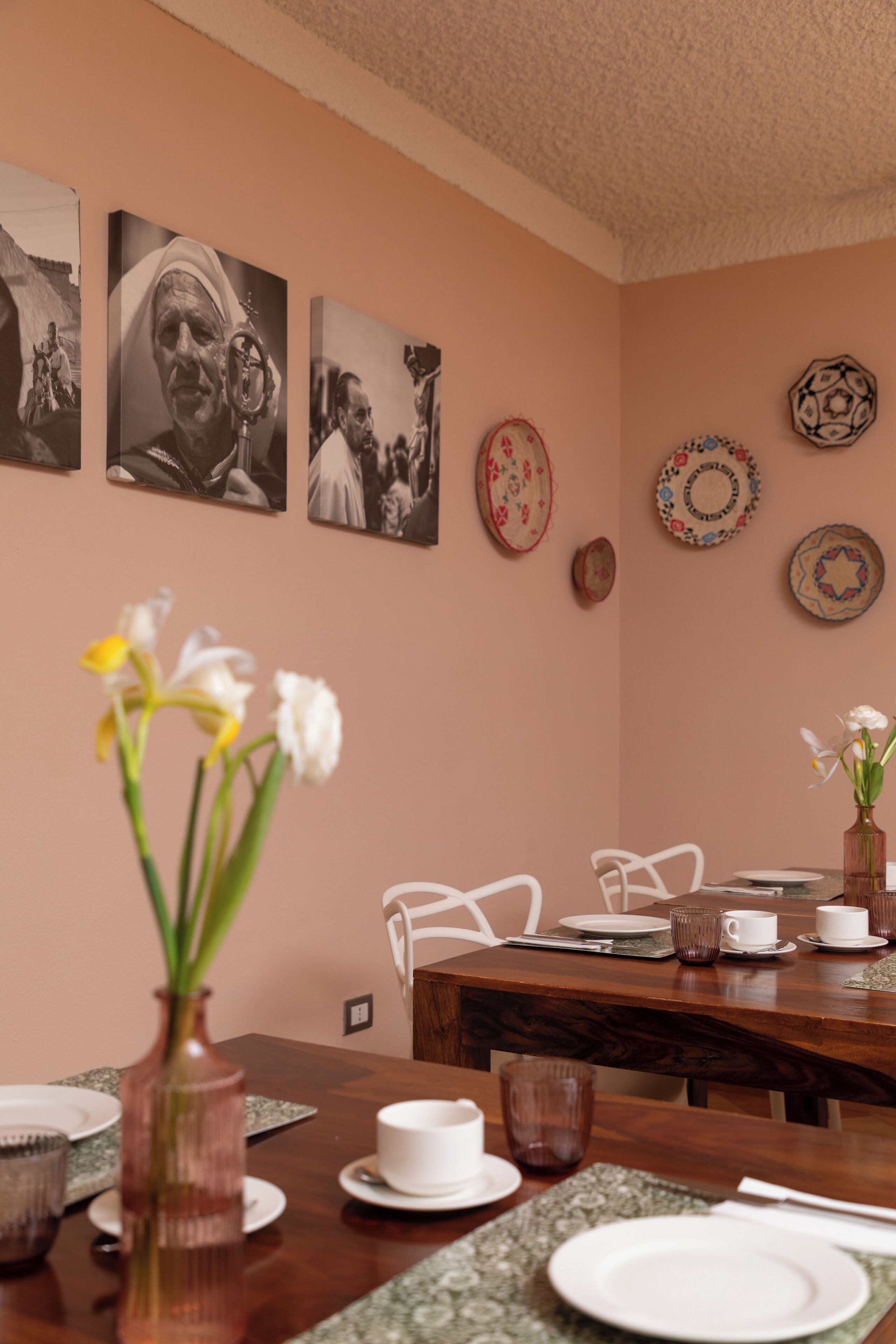A dining area with a wooden table, white chairs, a vase with flowers, and wall-mounted plates.