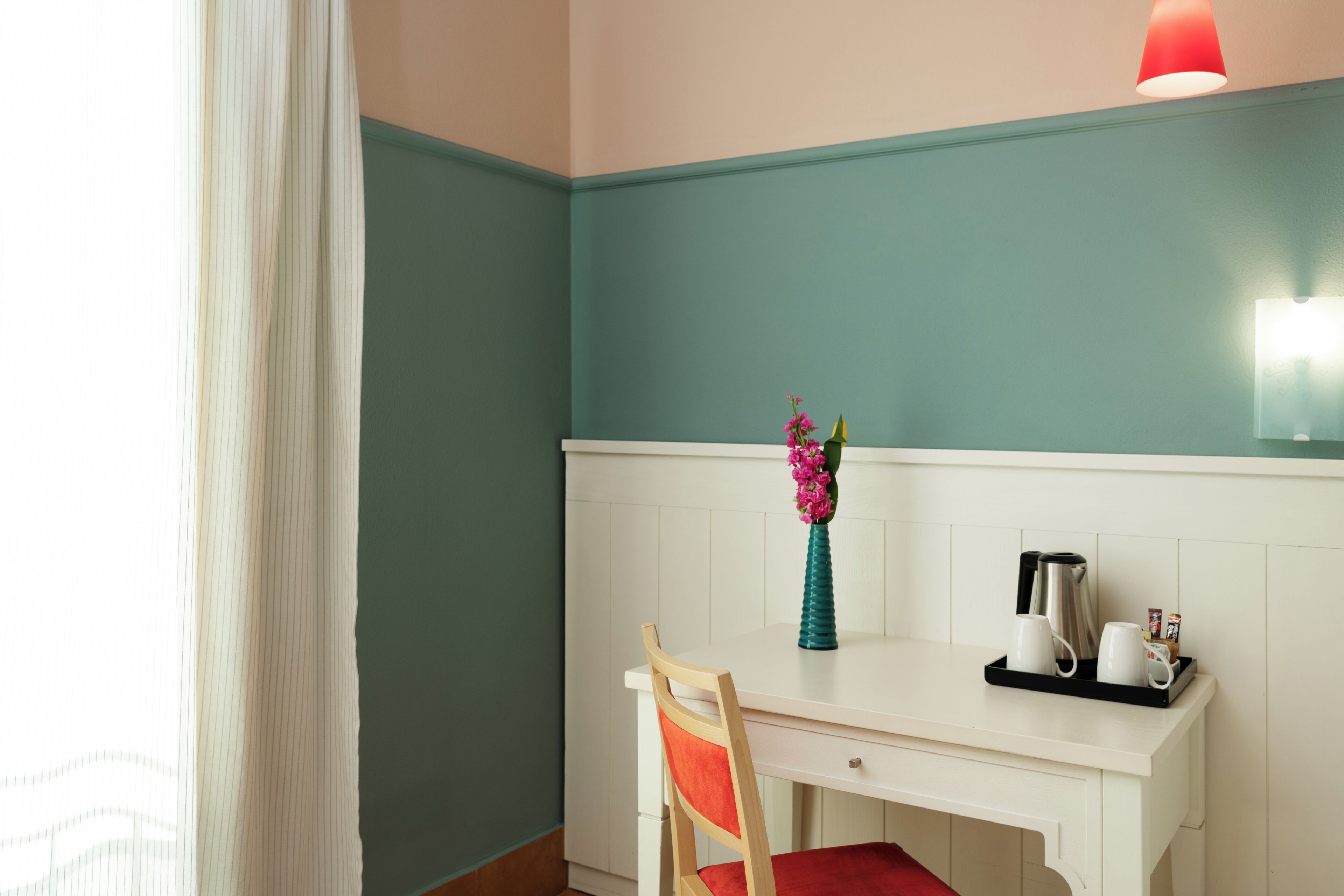 A room with a white desk, a chair, a vase with flowers, a tray with cups and a kettle, and a red light fixture.