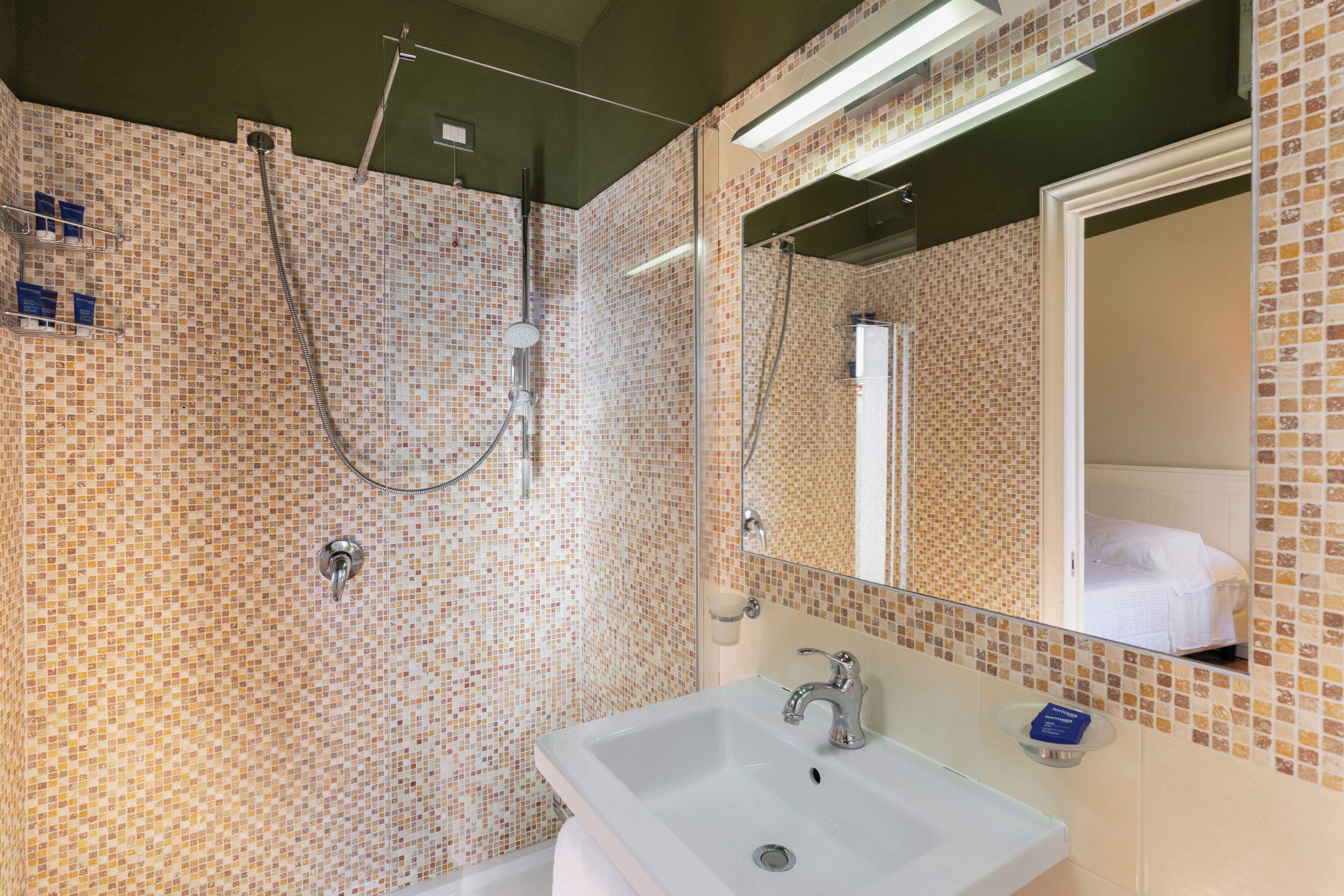 A bathroom with a tiled mosaic wall, a white sink, and a mirror reflecting a bed.