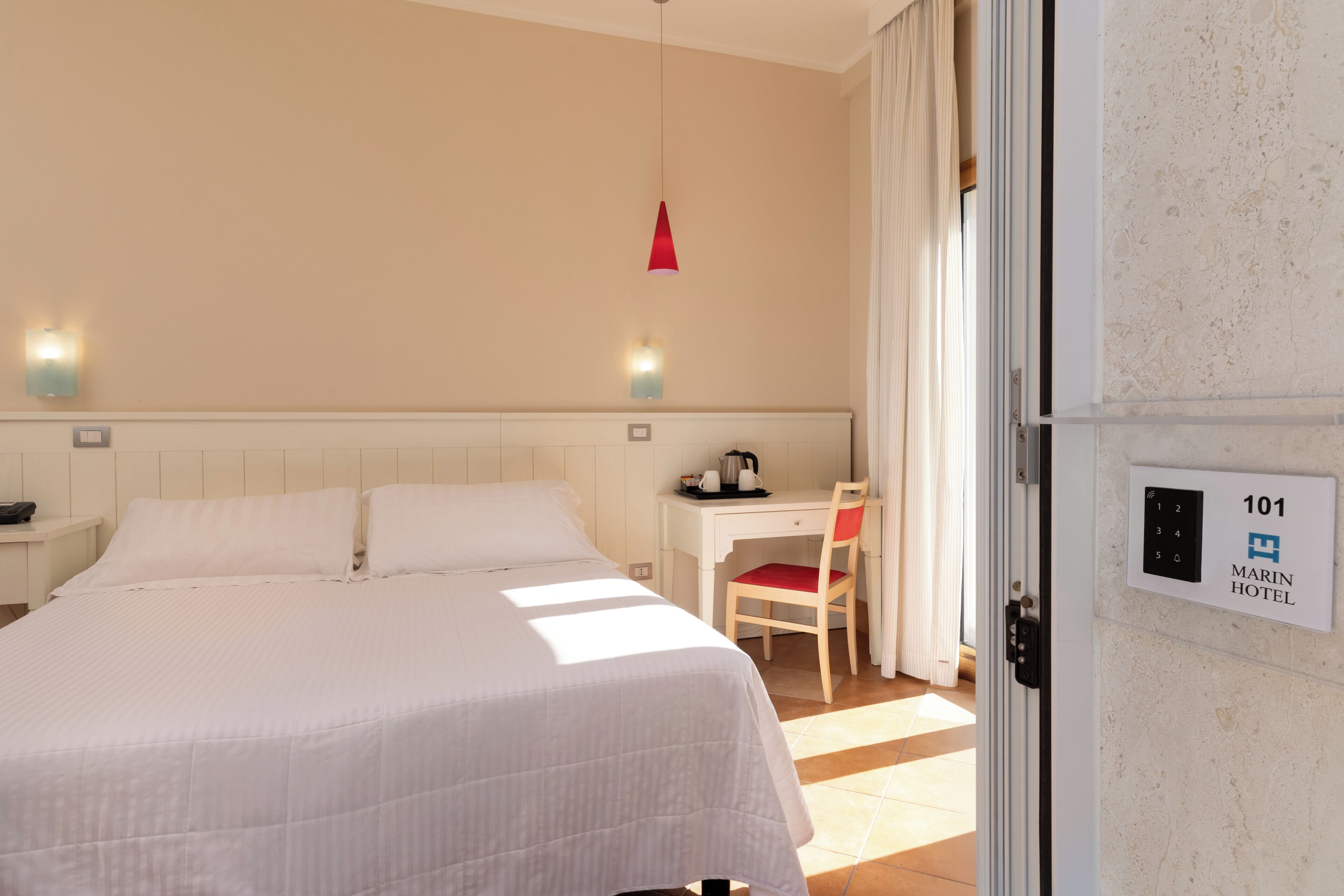 A hotel room with a bed, a desk, a chair, a red pendant light, and a door with a room number sign.
