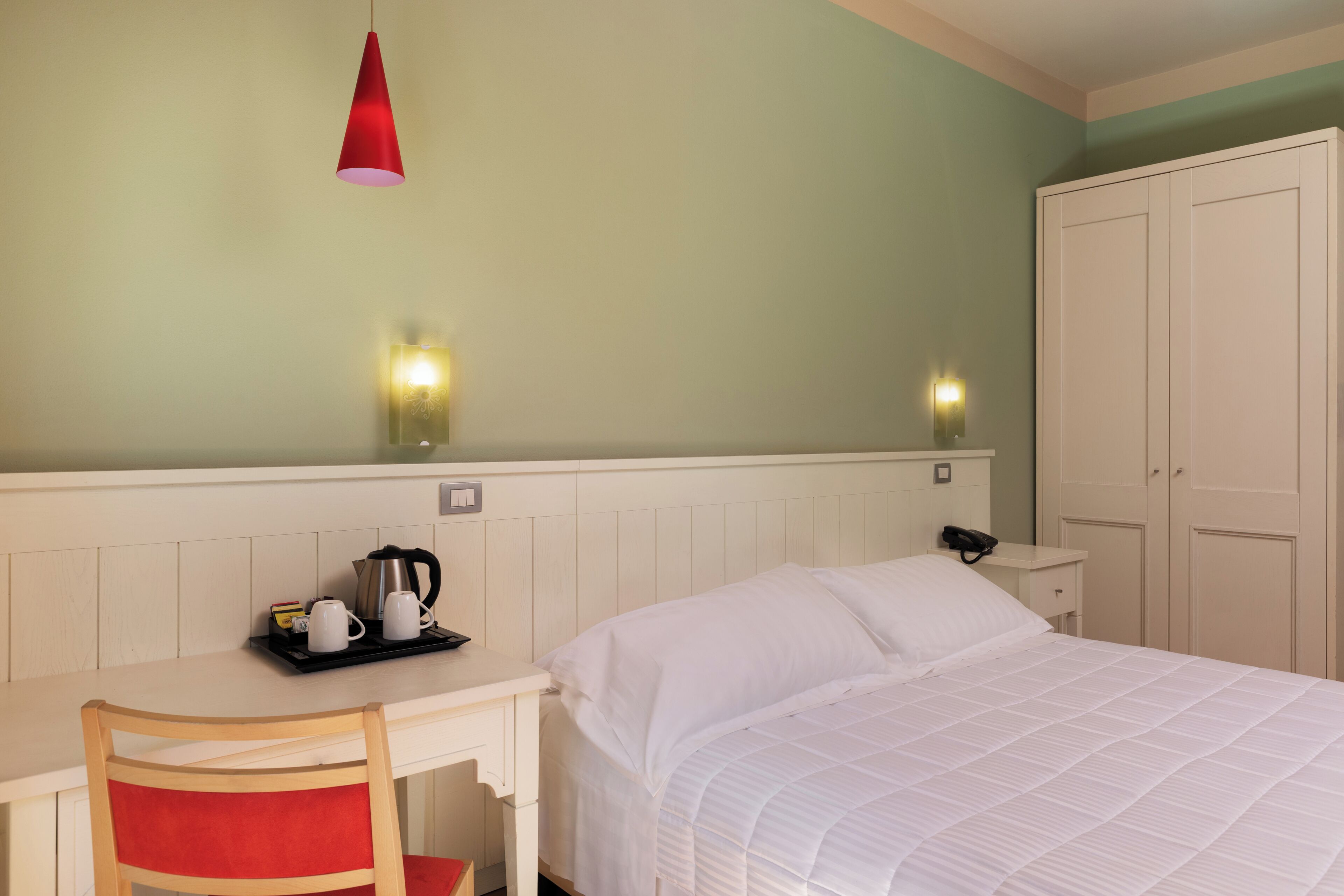 A hotel room with a bed, a desk with a chair, a red hanging light, and a wardrobe.