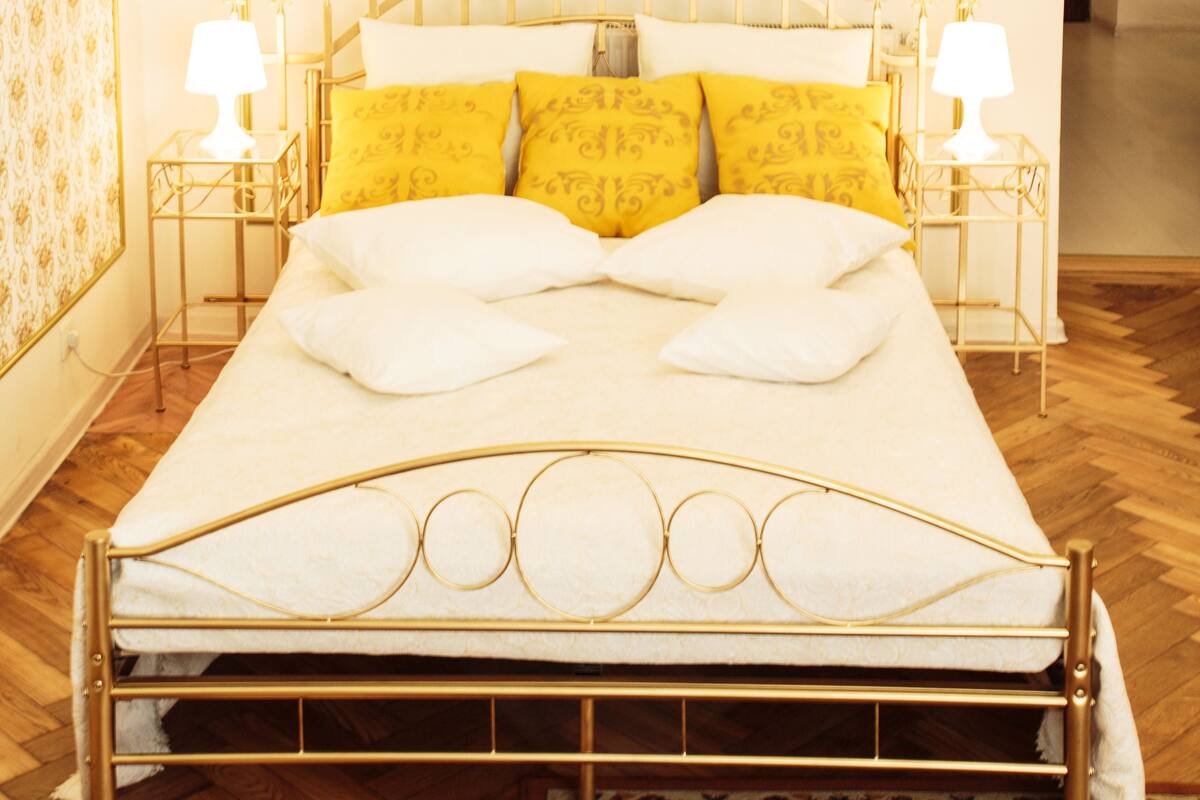 A bed with a gold frame, white bedding, and yellow pillows. There are two bedside tables with lamps.