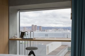 Een moderne hotelkamer met een groot raam met uitzicht op de stad, een houten bureau met een laptop en een klein krukje.