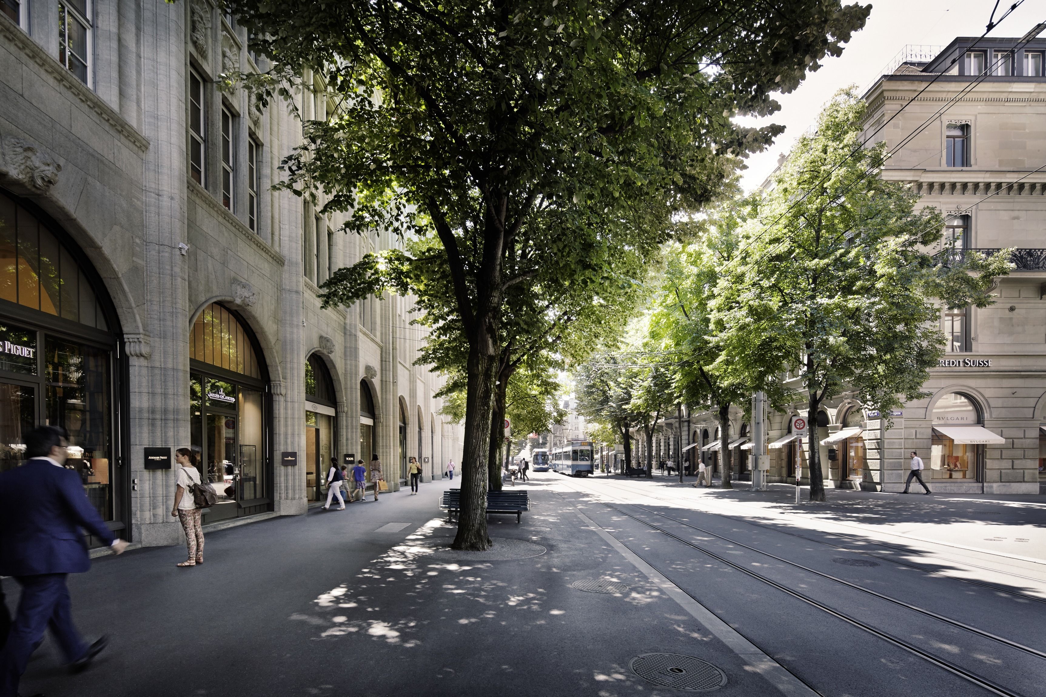 A tree-lined street with a tram, shops, and pedestrians.