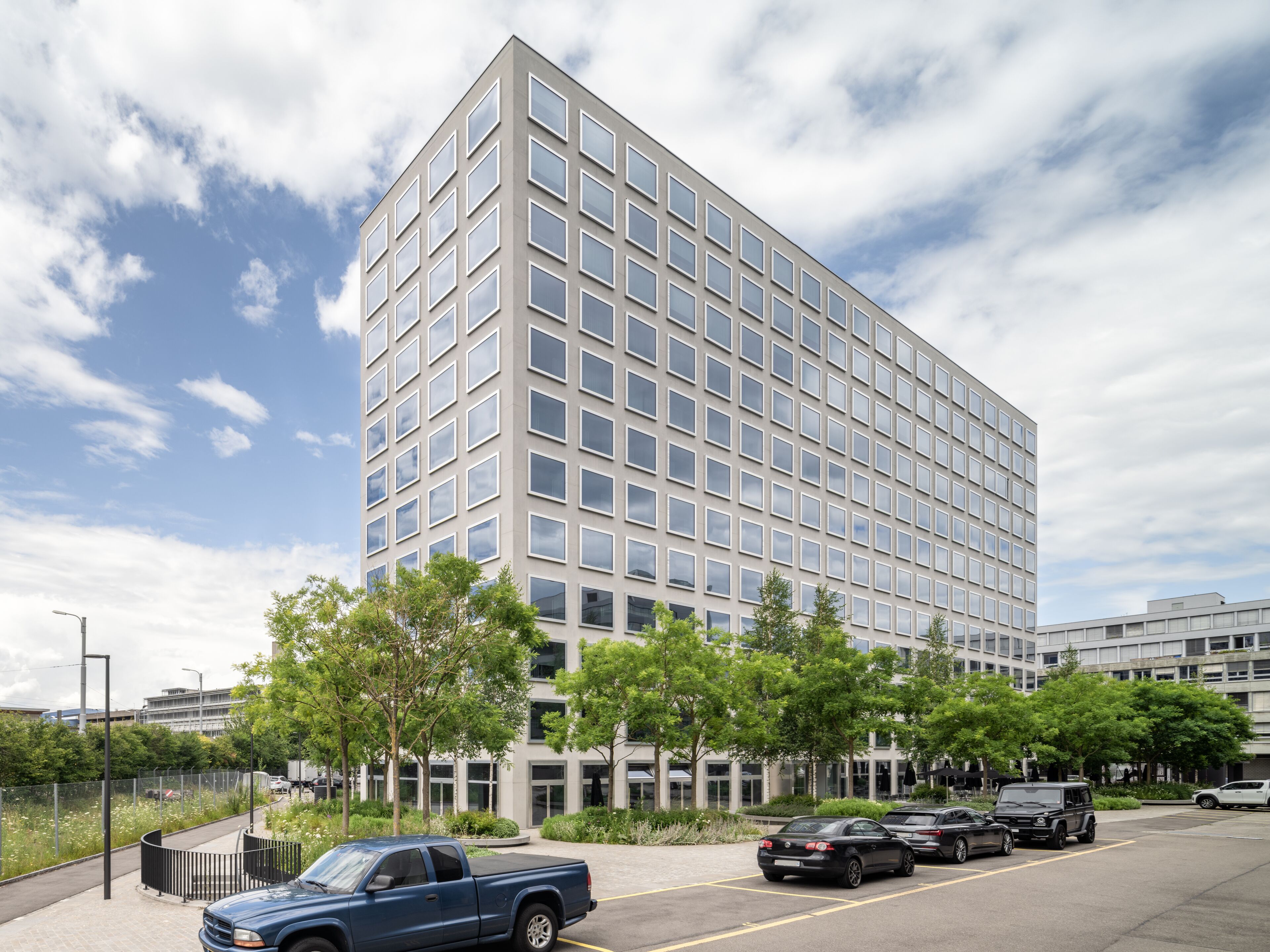 A modern building with a grid of windows, surrounded by trees and parked cars.