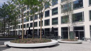 Een modern gebouw met een restaurant op de begane grond. Er zijn terrassen met tafels en stoelen, omgeven door bomen en plantenbakken.