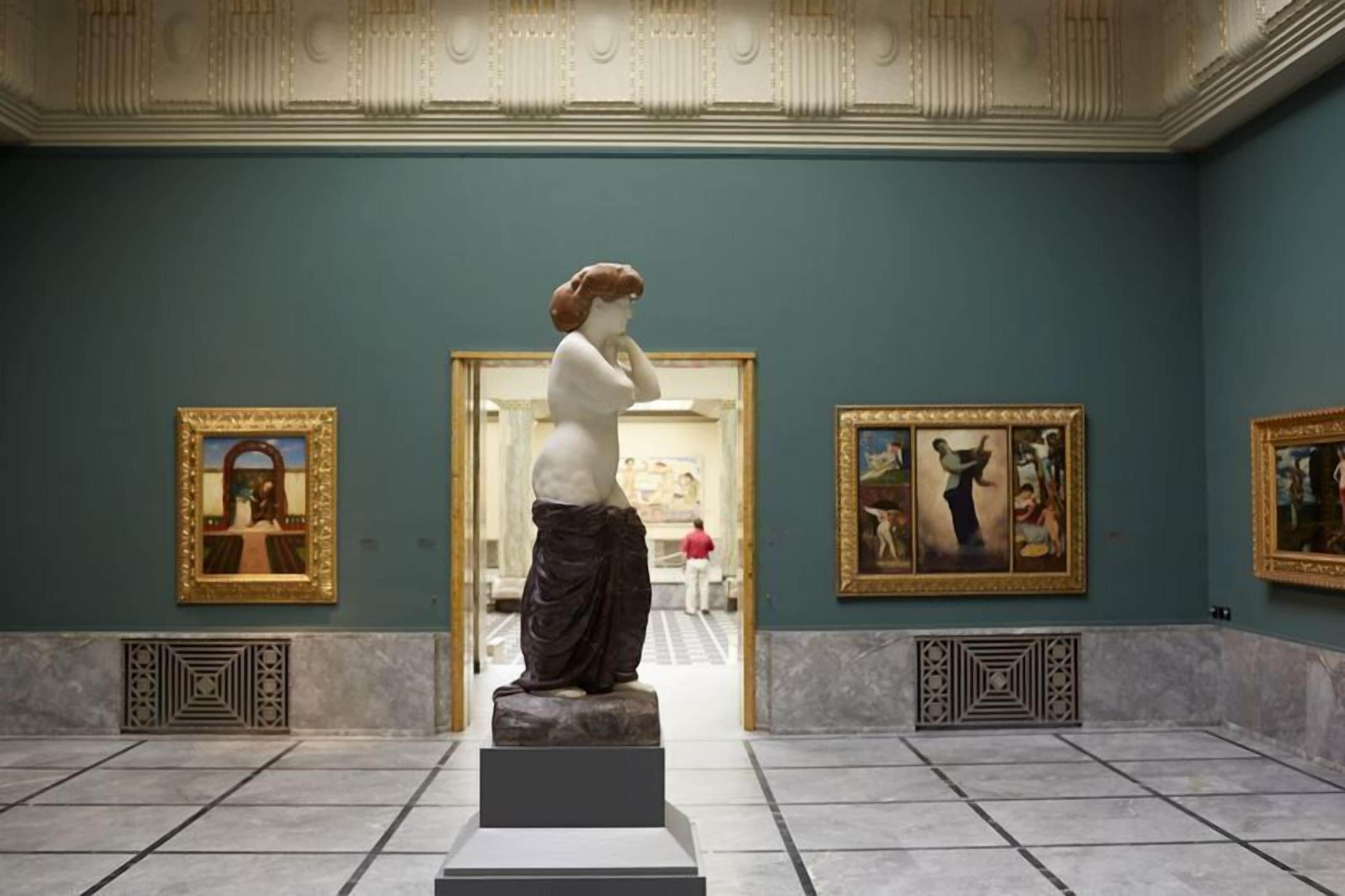 Ein Museuminnenraum mit einer zentralen Statue, gerahmten Gemälden und einem Fliesenboden.