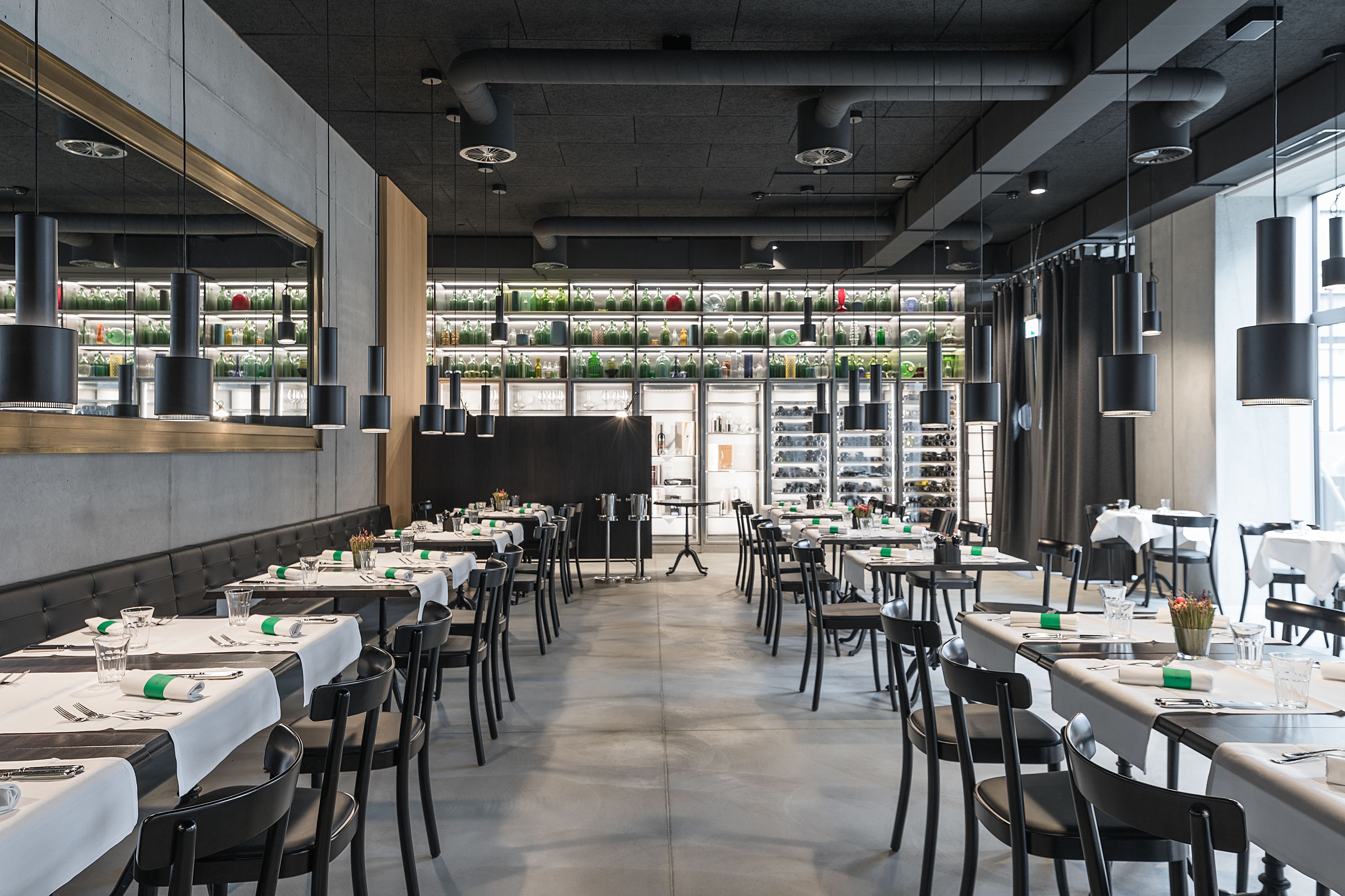 A restaurant with long tables, black chairs, and hanging pendant lights.