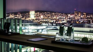 Une vue depuis une fenêtre donnant sur une ville illuminée la nuit, avec une table sur laquelle on aperçoit des verres, une tablette et des bouteilles d’eau.