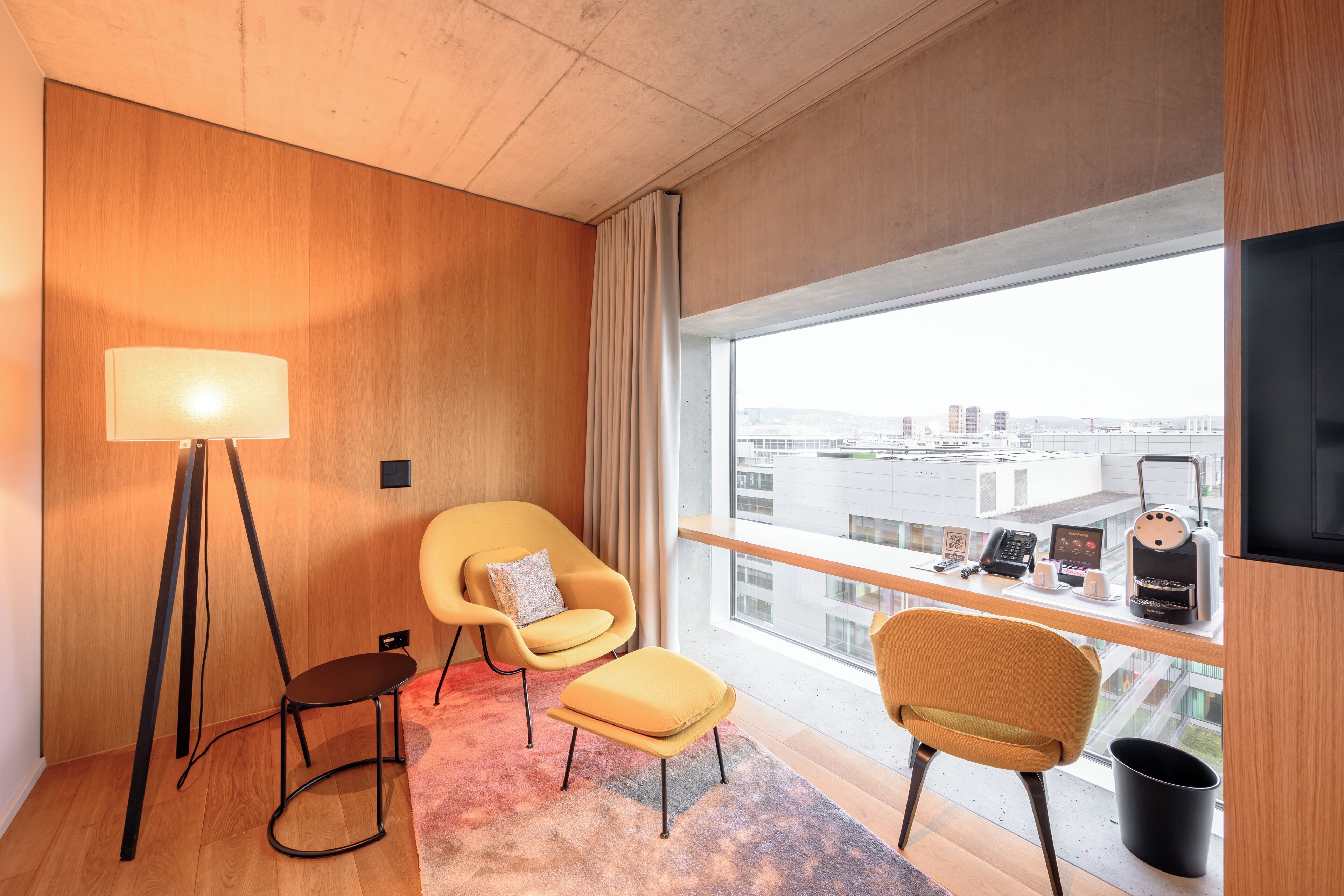 Habitación de hotel moderna con un ventanal grande, un escritorio con cafetera, una silla, una lámpara de pie y vistas a la ciudad.