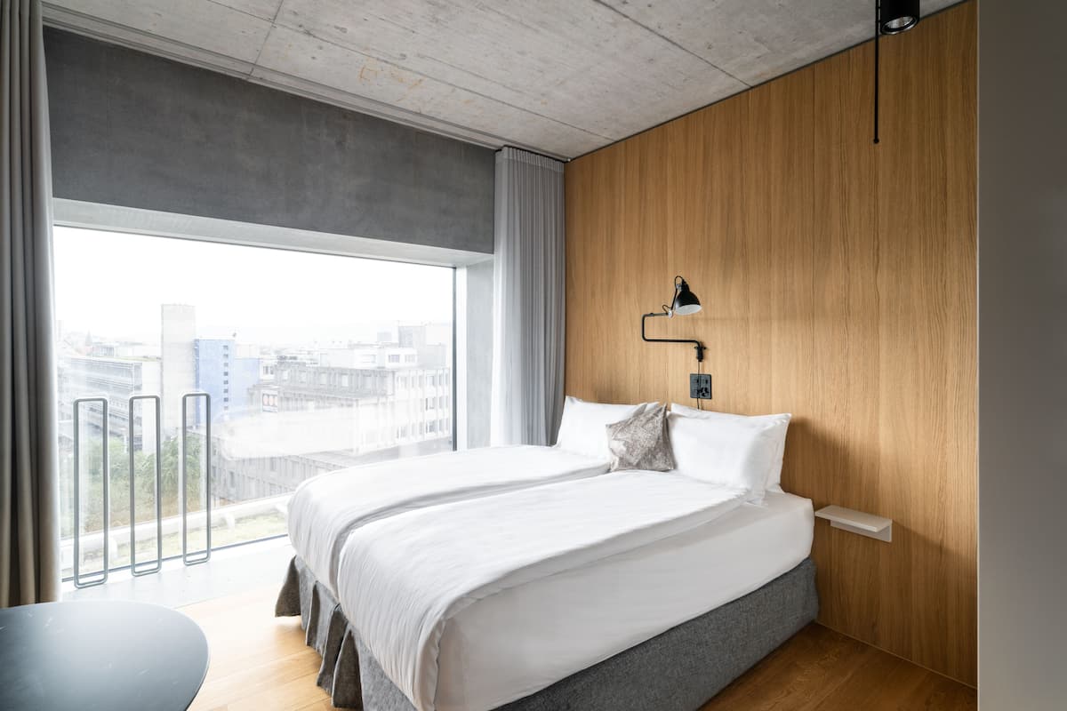 Une chambre d’hôtel moderne avec un grand lit, un mur décoratif en bois, une lampe de chevet et une vue sur la ville grâce à de grandes fenêtres.