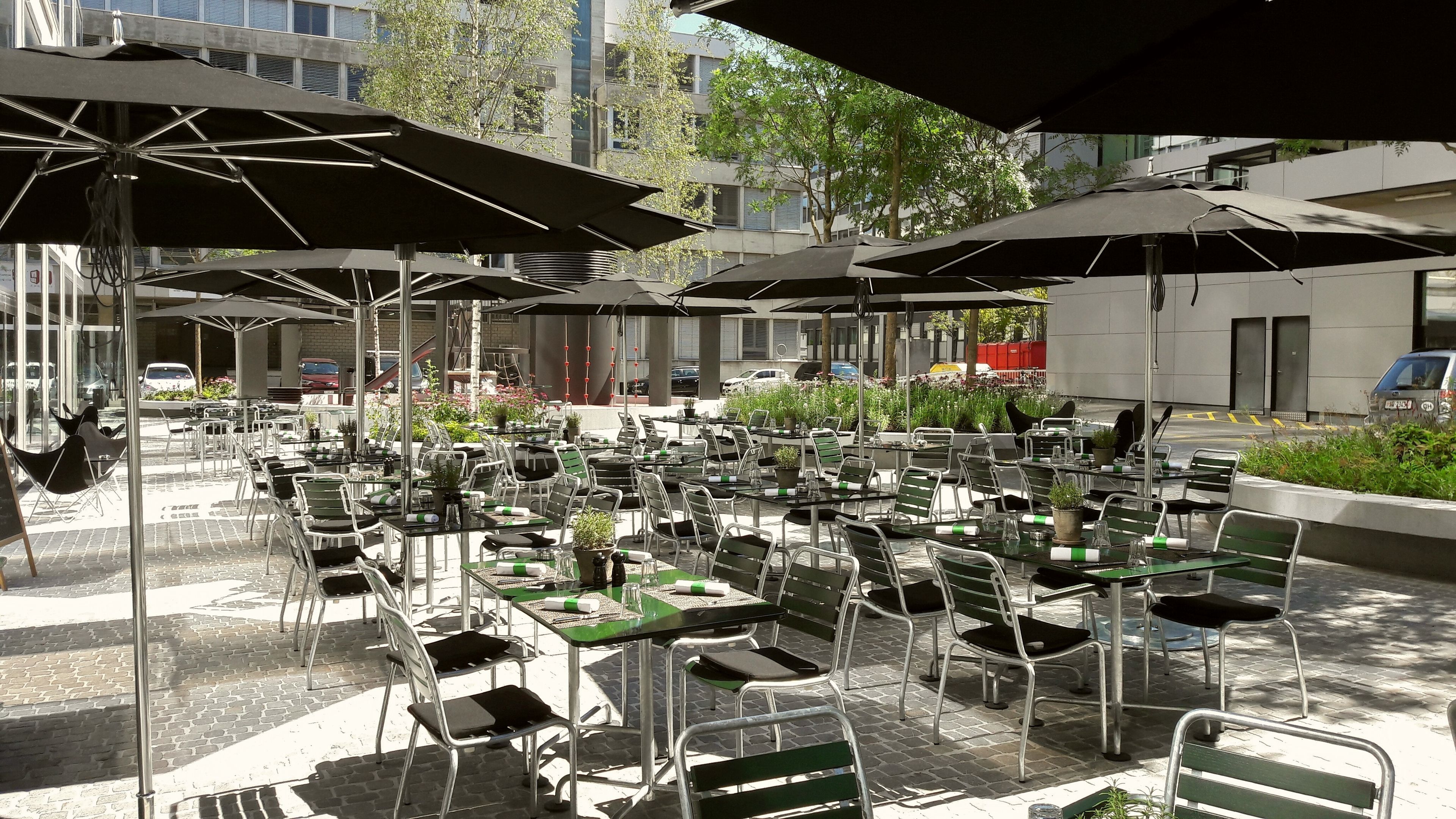 An outdoor café with multiple tables and chairs, shaded by umbrellas, set against a backdrop of buildings and trees.
