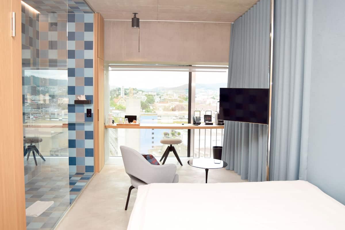 Une chambre d’hôtel moderne avec un grand lit, une chaise blanche, une petite table, un téléviseur et un balcon offrant une vue sur la ville.
