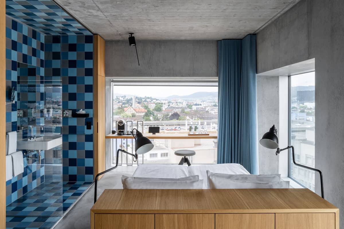 Une chambre d’hôtel moderne dotée d’une grande fenêtre, d’une table de chevet en bois, d’un bureau avec un ordinateur et offrant une vue sur le paysage urbain.